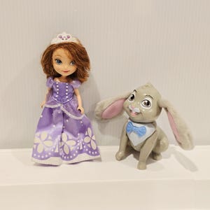 May include: A small doll with brown hair wearing a purple dress with white floral patterns and a white crown, standing next to a gray rabbit figurine with long ears, a white chest patch, and a blue bow tie. Both figures are standing on a white surface.