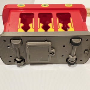 K&ouml;nnte beinhalten: Ein roter und gelber Spielzeugbus mit grauem Sockel, mit vier gelben Sitzen, die durch die Fenster sichtbar sind. Der Bus hat vier schwarze R&auml;der und ein Batteriefach an der Unterseite. Die Oberseite des Busses hat drei gr&uuml;ne Kn&ouml;pfe.