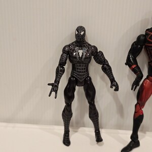 May include: A black Spider-Man action figure with a white spider emblem on the chest. The figure has a detailed web pattern and is posed with arms outstretched. The figure is approximately 6 inches tall.