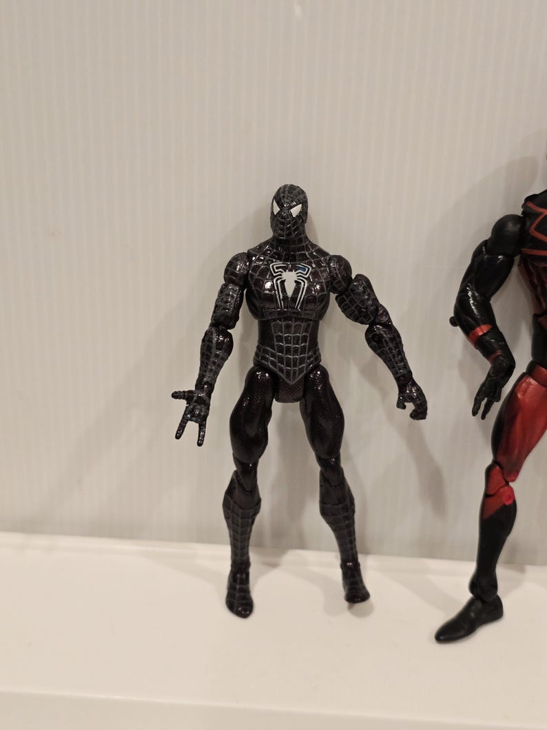May include: A black Spider-Man action figure with a white spider emblem on the chest. The figure has a detailed web pattern and is posed with arms outstretched. The figure is approximately 6 inches tall.