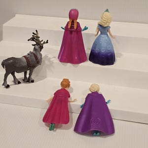 May include: A collection of toy figurines featuring characters from the animated film "Frozen." The set includes Anna, Elsa, and a reindeer. The figures are brightly coloured and made of plastic.