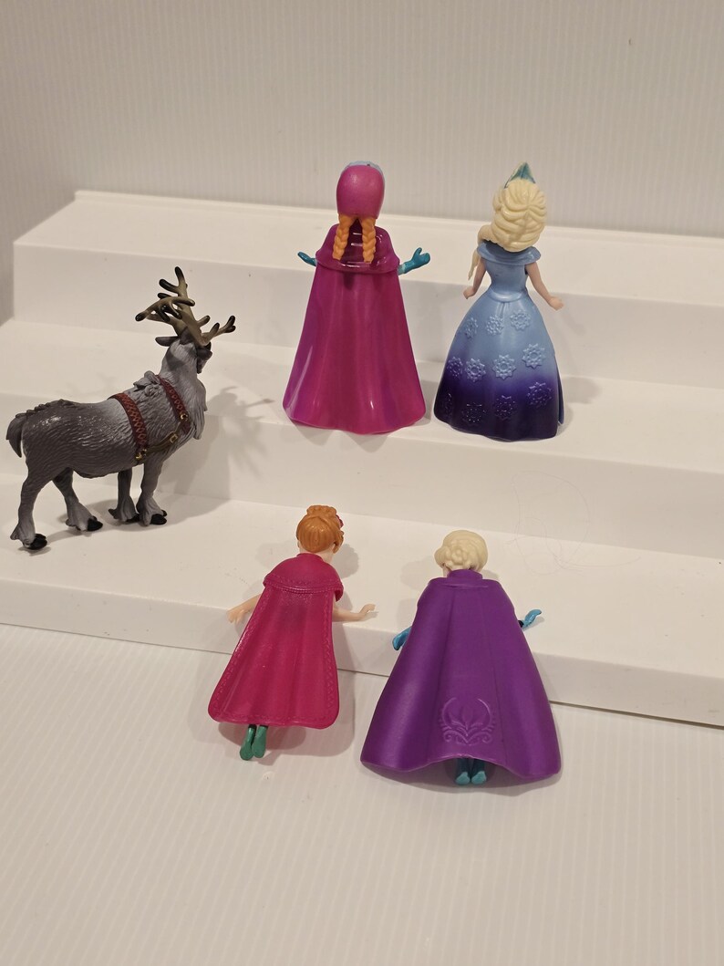 May include: A collection of toy figurines featuring characters from the animated film "Frozen." The set includes Anna, Elsa, and a reindeer. The figures are brightly coloured and made of plastic.