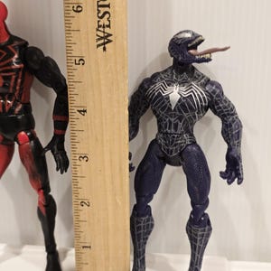 May include: A Venom action figure, approximately 6 inches tall, stands next to a wooden ruler. The figure is primarily dark blue with white spiderweb detailing and a white spider emblem. Its tongue is extended.