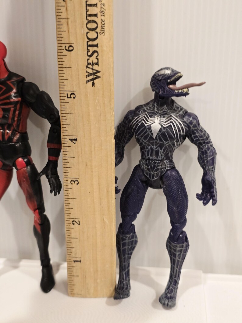 May include: A Venom action figure, approximately 6 inches tall, stands next to a wooden ruler. The figure is primarily dark blue with white spiderweb detailing and a white spider emblem. Its tongue is extended.