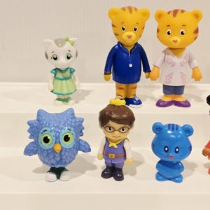 May include: A collection of six small plastic character figurines including a white cat in a green dress, two orange tigers in blue and white outfits, a blue owl, a human figure with brown hair and glasses, and a small blue cat-like creature.
