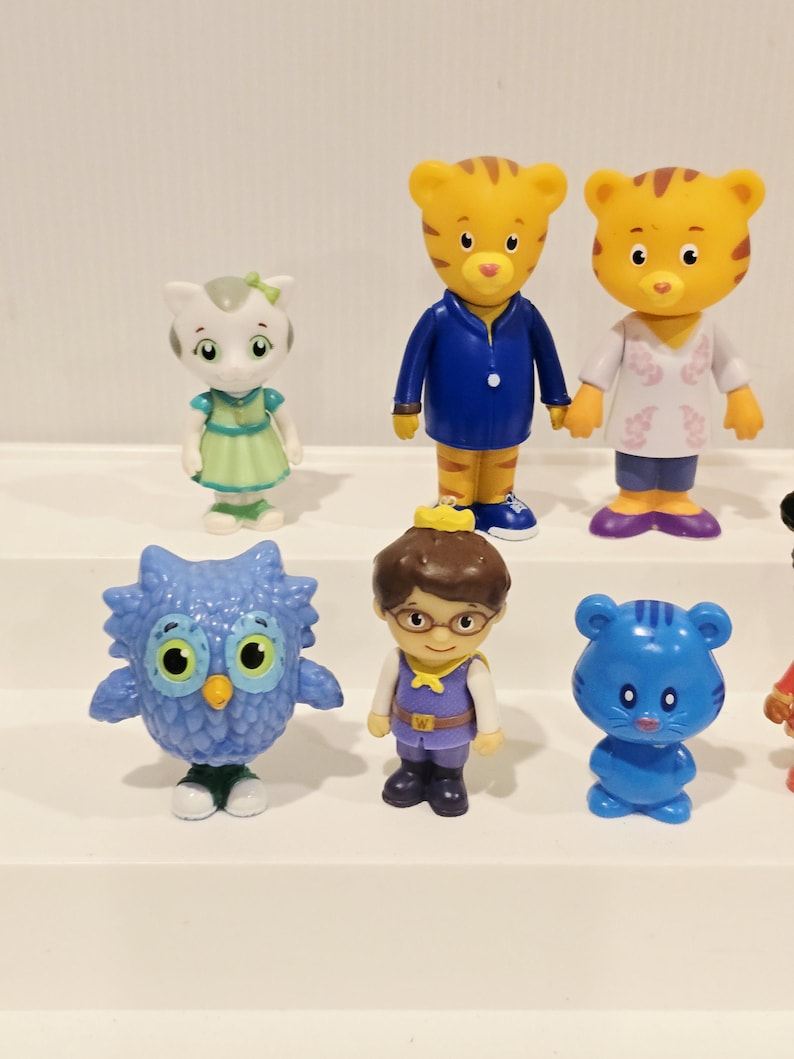 May include: A collection of six small plastic character figurines including a white cat in a green dress, two orange tigers in blue and white outfits, a blue owl, a human figure with brown hair and glasses, and a small blue cat-like creature.