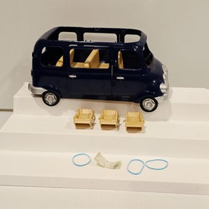 May include: A dark blue toy van with a light tan interior and three matching chairs. The van has silver-colored wheels and trim. Two blue rubber bands and a small piece of paper are also present.