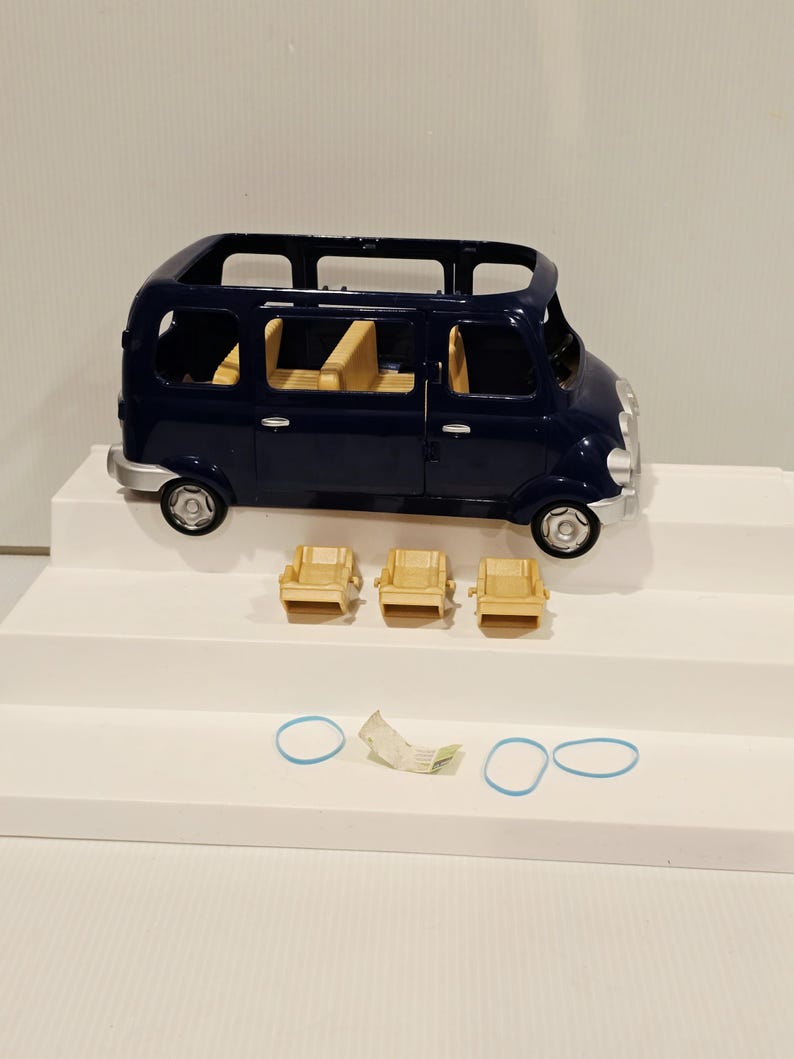 May include: A dark blue toy van with a light tan interior and three matching chairs. The van has silver-colored wheels and trim. Two blue rubber bands and a small piece of paper are also present.