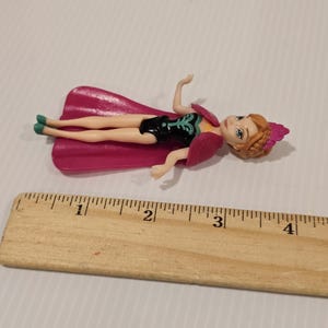 May include: A small doll of a character from the movie Frozen, lying on its back. The doll is wearing a black dress with a teal design, a pink cape, and a pink headpiece. The doll is approximately 7.6 cm long.