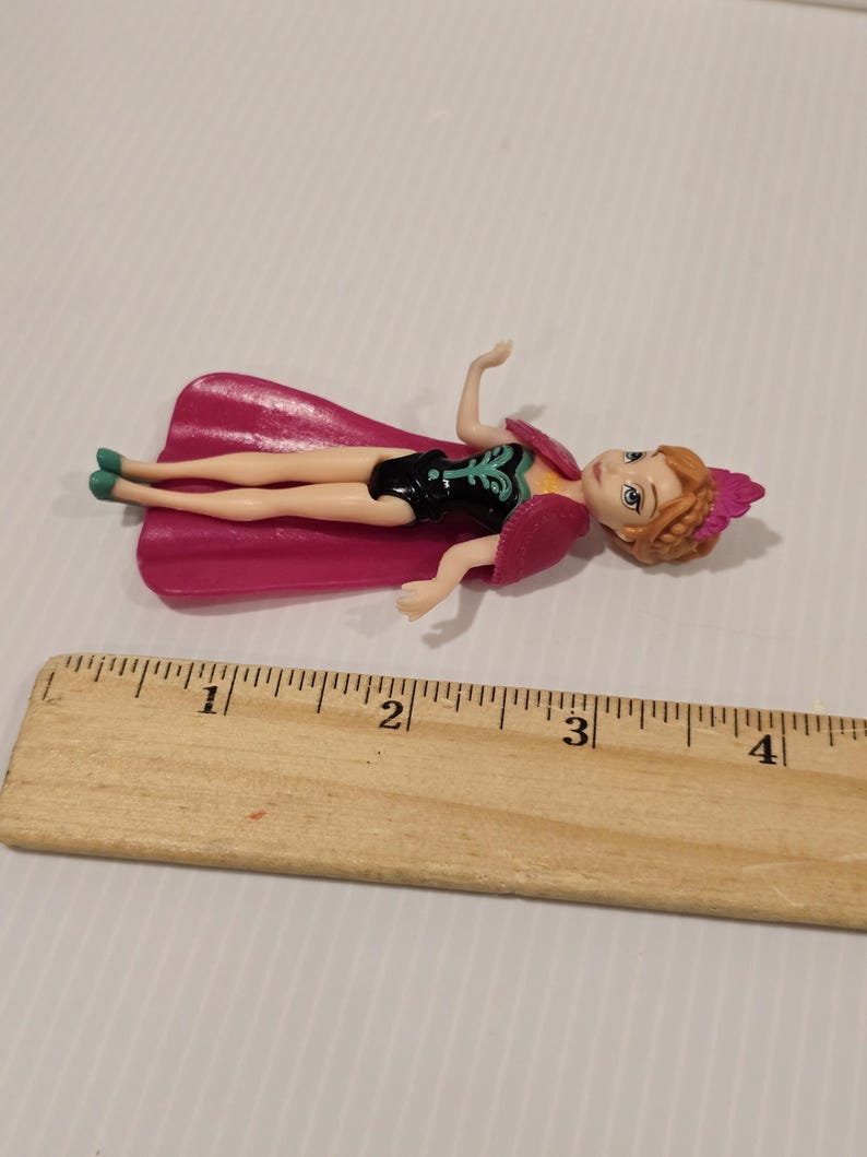 May include: A small doll of a character from the movie Frozen, lying on its back. The doll is wearing a black dress with a teal design, a pink cape, and a pink headpiece. The doll is approximately 7.6 cm long.