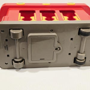 K&ouml;nnte beinhalten: Ein roter und gelber Spielzeugbus mit grauem Sockel, R&auml;dern und einem Batteriefach. Der Bus hat einen Schalter mit der Aufschrift "TRY ME OFF ON".