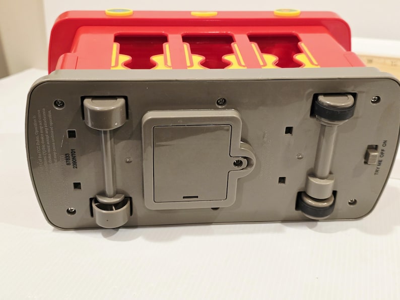 K&ouml;nnte beinhalten: Ein roter und gelber Spielzeugbus mit grauem Sockel, R&auml;dern und einem Batteriefach. Der Bus hat einen Schalter mit der Aufschrift "TRY ME OFF ON".