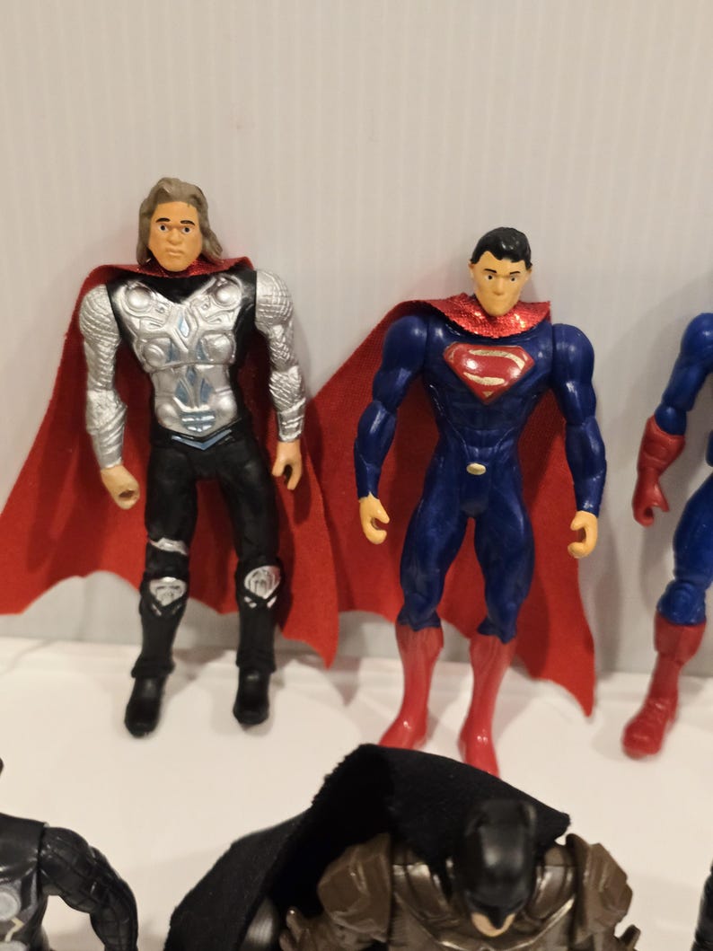May include: A collection of superhero action figures, including Thor and Superman. Thor has long blonde hair, a silver chest plate, and a red cape. Superman wears a blue suit with a red cape and boots. Other figures are partially visible.