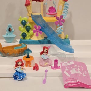 K&ouml;nnte beinhalten: Ariel's Sea Castle Spielset mit einem gelb, rosa und blau gef&auml;rbten Schloss, einer Rutsche und einer rosa Oktopus-Schaukel. Das Set enth&auml;lt Ariel-Figuren, ein Boot, einen Tisch und weiteres Zubeh&ouml;r. Die Verpackung tr&auml;gt die Aufschrift "Ariel's Sea Castle".