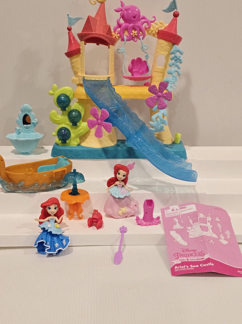 K&ouml;nnte beinhalten: Ariel's Sea Castle Spielset mit einem gelb, rosa und blau gef&auml;rbten Schloss, einer Rutsche und einer rosa Oktopus-Schaukel. Das Set enth&auml;lt Ariel-Figuren, ein Boot, einen Tisch und weiteres Zubeh&ouml;r. Die Verpackung tr&auml;gt die Aufschrift "Ariel's Sea Castle".