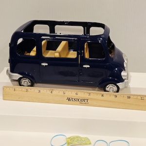 May include: A dark blue toy van with yellow interior seating. The van has silver accents and black wheels. A wooden ruler with the text "WESTCOTT Since 1872" is next to the van. Two blue rubber bands and a small green and yellow paper are also present.