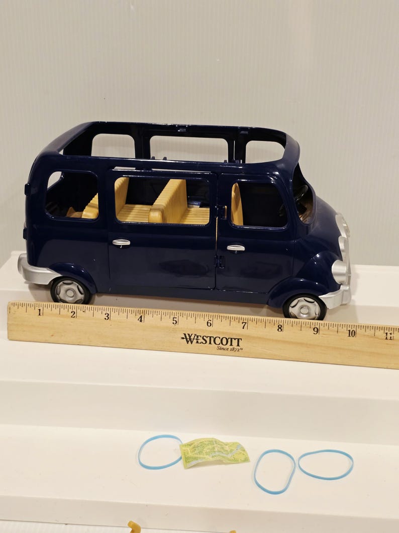 May include: A dark blue toy van with yellow interior seating. The van has silver accents and black wheels. A wooden ruler with the text "WESTCOTT Since 1872" is next to the van. Two blue rubber bands and a small green and yellow paper are also present.