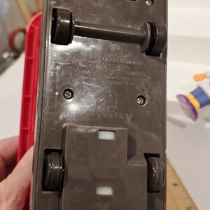 May include: Underside view of a brown toy vehicle with black wheels and axles. The toy has a rectangular base with visible screws and the text "The Fred Rogers Company" and "Made in China".