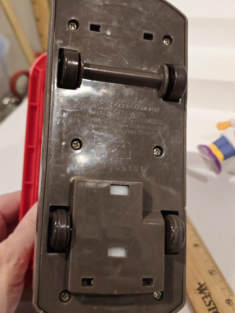 May include: Underside view of a brown toy vehicle with black wheels and axles. The toy has a rectangular base with visible screws and the text "The Fred Rogers Company" and "Made in China".