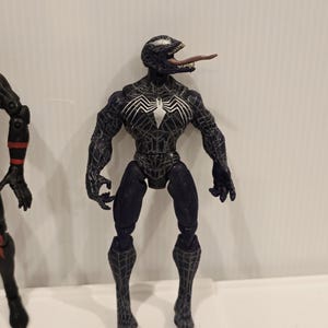 May include: A black Venom action figure with a white spider emblem on the chest and a long, pink tongue. The figure has a detailed, textured design and is posed standing upright. The figure is a collectible item.