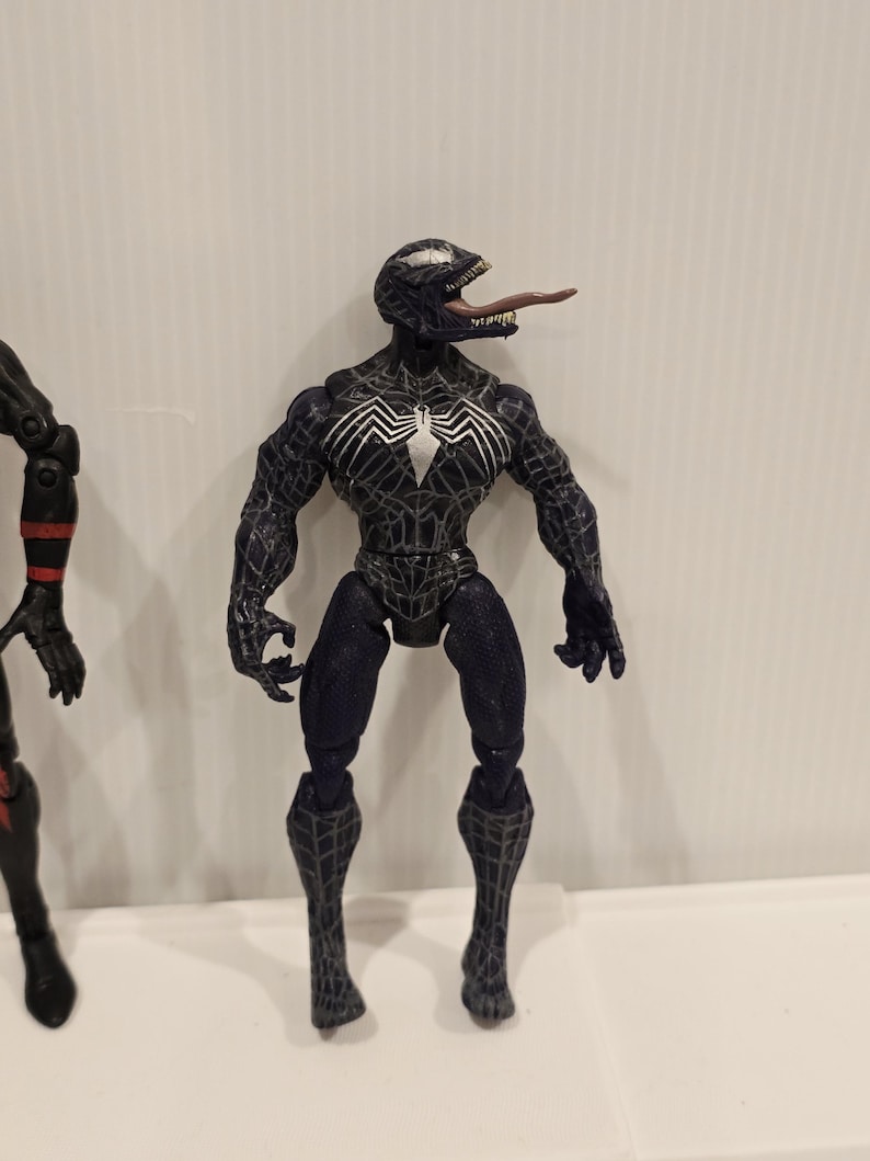 May include: A black Venom action figure with a white spider emblem on the chest and a long, pink tongue. The figure has a detailed, textured design and is posed standing upright. The figure is a collectible item.