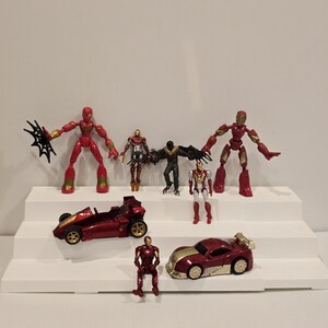 May include: A collection of red and gold action figures and toy cars, including Iron Man and Spider-Man characters. The toys are arranged on a white tiered display, showcasing various poses and vehicle designs.
