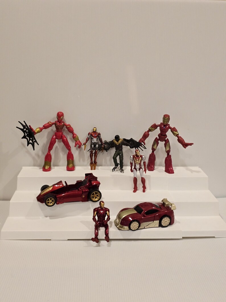 May include: A collection of red and gold action figures and toy cars, including Iron Man and Spider-Man characters. The toys are arranged on a white tiered display, showcasing various poses and vehicle designs.
