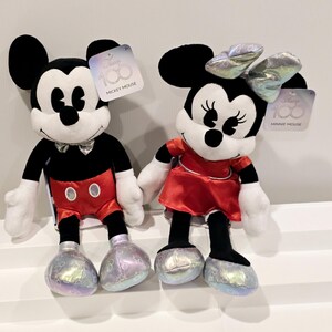 以下が含まれることがあります： ミッキーマウスとミニーマウスのぬいぐるみが、クラシックな黒と白と赤の衣装を着ています。どちらもキラキラした仕上げの銀色の靴を履いています。おもちゃはミッキーマウスの100周年を祝っています。