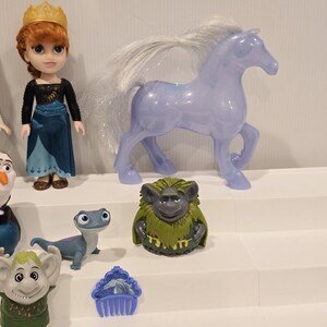 May include: A collection of Frozen-themed toys, including a doll with a gold crown, a translucent purple horse, and various character figurines. The toys are arranged on a white tiered display.
