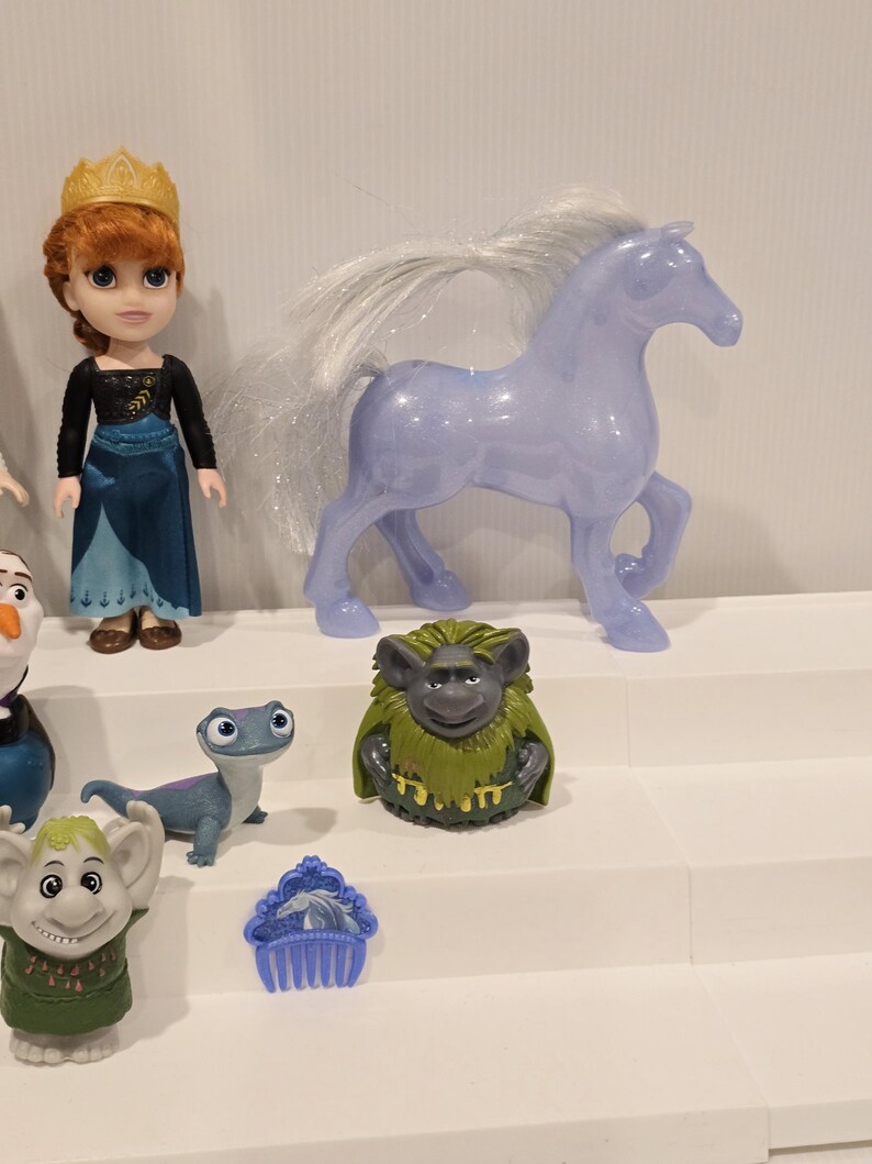 May include: A collection of Frozen-themed toys, including a doll with a gold crown, a translucent purple horse, and various character figurines. The toys are arranged on a white tiered display.