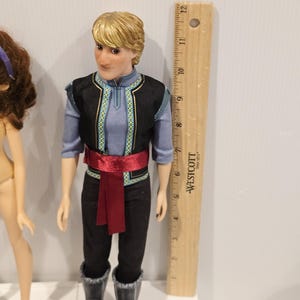 May include: A doll of a male character with blonde hair, wearing a blue shirt, black vest with gold trim, black trousers, and black boots. A red sash is tied around the waist. A wooden ruler is next to the doll, measuring approximately 30 cm.