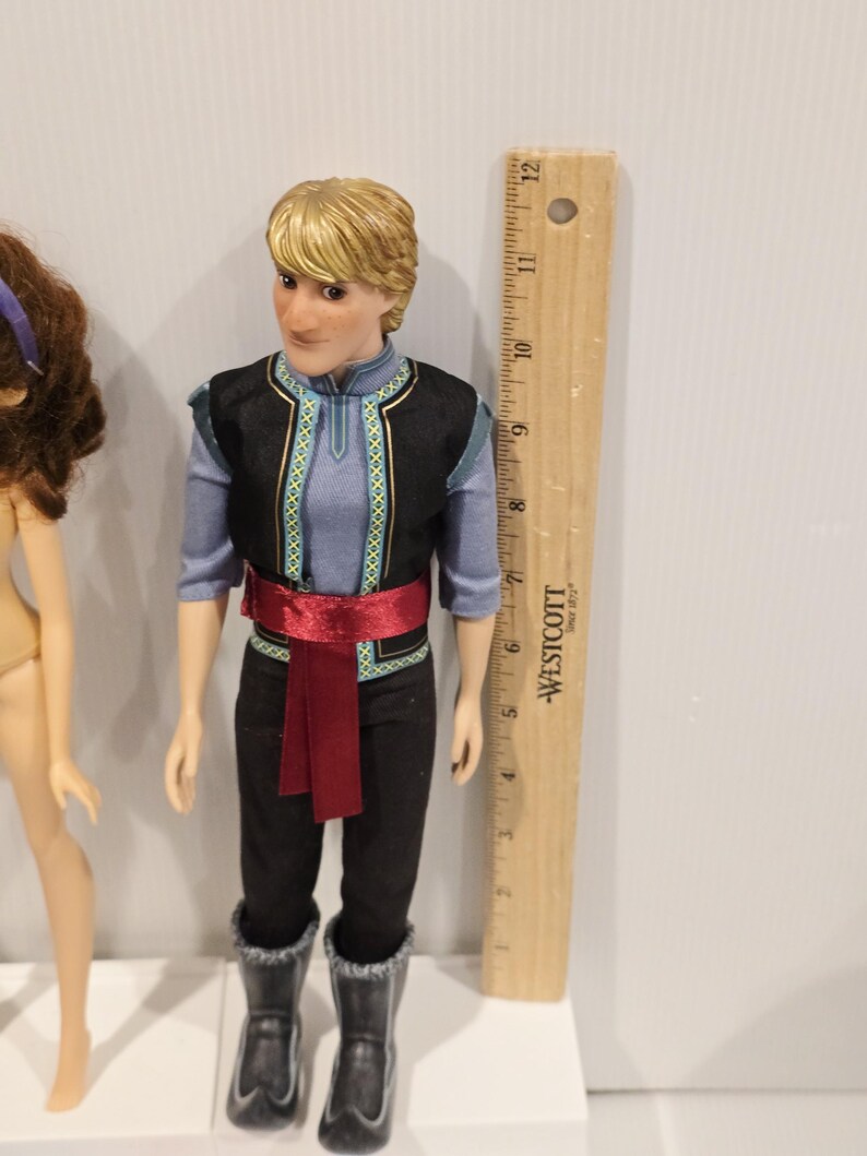 May include: A doll of a male character with blonde hair, wearing a blue shirt, black vest with gold trim, black trousers, and black boots. A red sash is tied around the waist. A wooden ruler is next to the doll, measuring approximately 30 cm.