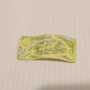 May include: A small, folded map with a detailed illustration of a rural landscape. The map features green trees, blue rivers, and small buildings, suggesting a countryside or village setting. The map is on a light green background.