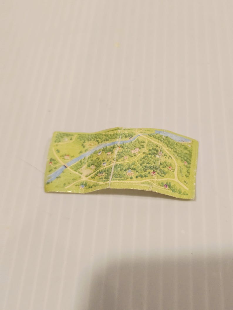 May include: A small, folded map with a detailed illustration of a rural landscape. The map features green trees, blue rivers, and small buildings, suggesting a countryside or village setting. The map is on a light green background.