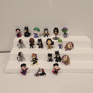 May include: A collection of small superhero figurines, including Batman, Superman, and others. The figures are brightly colored and posed on a white stepped display. The collection features various characters from the DC Comics universe.