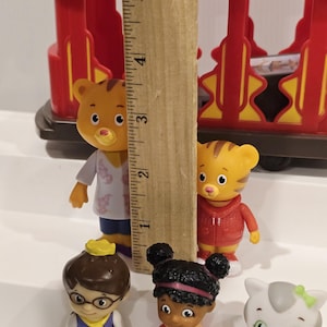 May include: A collection of Daniel Tiger's Neighborhood figurines, including Daniel Tiger, Katerina Kittycat, and O the Owl. The figurines are various colors and stand in front of a red toy train car. A wooden ruler is in the center.