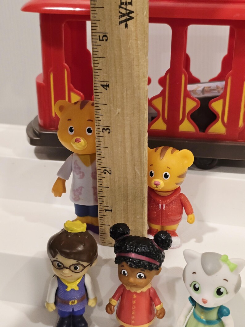 May include: A collection of Daniel Tiger's Neighborhood figurines, including Daniel Tiger, Katerina Kittycat, and O the Owl. The figurines are various colors and stand in front of a red toy train car. A wooden ruler is in the center.