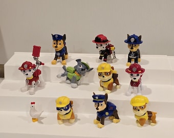May include: A collection of colorful Paw Patrol character figurines. The figurines include various dog characters in uniforms and hats, along with a small white chicken figurine. The toys are arranged on a white stepped display.