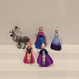 May include: A selection of miniature figurines depicting characters from the animated film "Frozen." The set includes Elsa, Anna, and other characters in their iconic outfits. A reindeer figurine is also present. The figurines are made of plastic and are brightly coloured.
