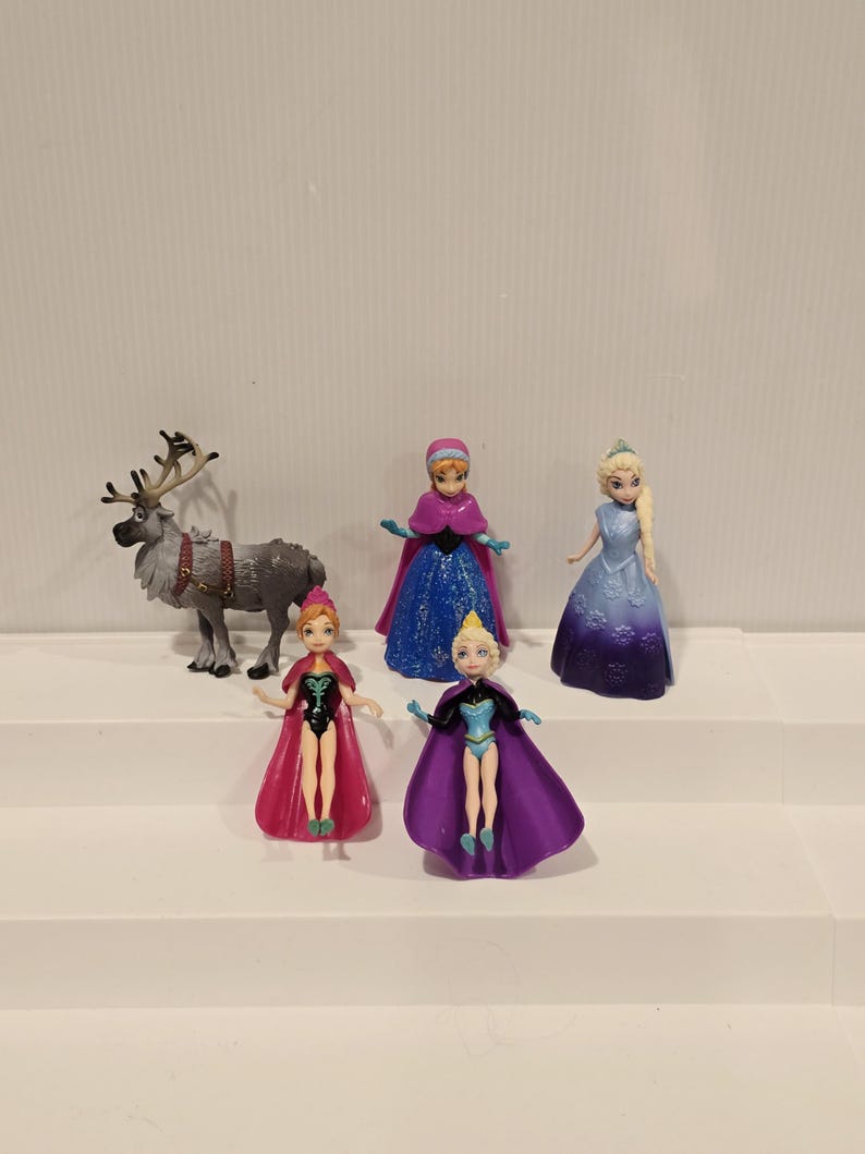 May include: A selection of miniature figurines depicting characters from the animated film "Frozen." The set includes Elsa, Anna, and other characters in their iconic outfits. A reindeer figurine is also present. The figurines are made of plastic and are brightly coloured.