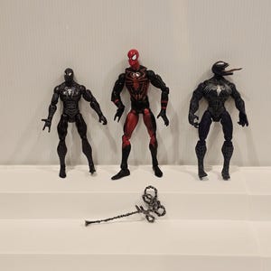 May include: Three action figures of Spider-Man and Venom characters. The Spider-Man figures are in black and red, and the Venom figure is black and blue. A silver web shooter is in front of the figures.