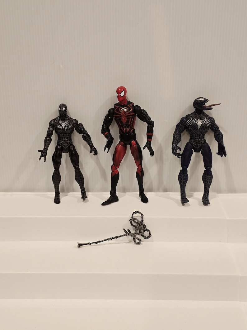 May include: Three action figures of Spider-Man and Venom characters. The Spider-Man figures are in black and red, and the Venom figure is black and blue. A silver web shooter is in front of the figures.