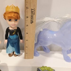 May include: A doll of a character from the movie Frozen, wearing a blue and black dress and a gold crown. A light blue toy horse with a white mane is next to the doll. A wooden ruler is in the background.