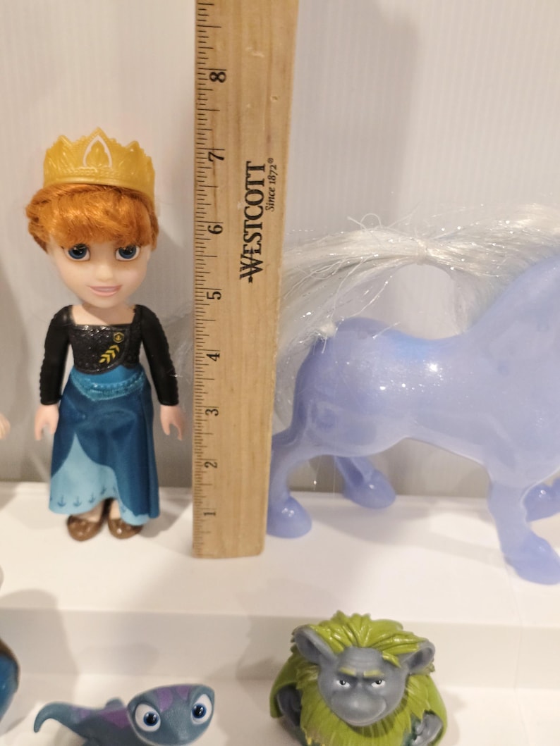 May include: A doll of a character from the movie Frozen, wearing a blue and black dress and a gold crown. A light blue toy horse with a white mane is next to the doll. A wooden ruler is in the background.