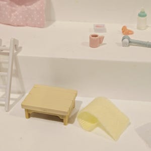 Puede incluir: Una escena en miniatura con una peque&ntilde;a mesa de picnic de color amarillo claro con una parte superior de listones y una toalla amarilla enrollada. Otros art&iacute;culos incluyen una taza rosa, un biber&oacute;n y una peque&ntilde;a almohada rosa con lunares blancos.