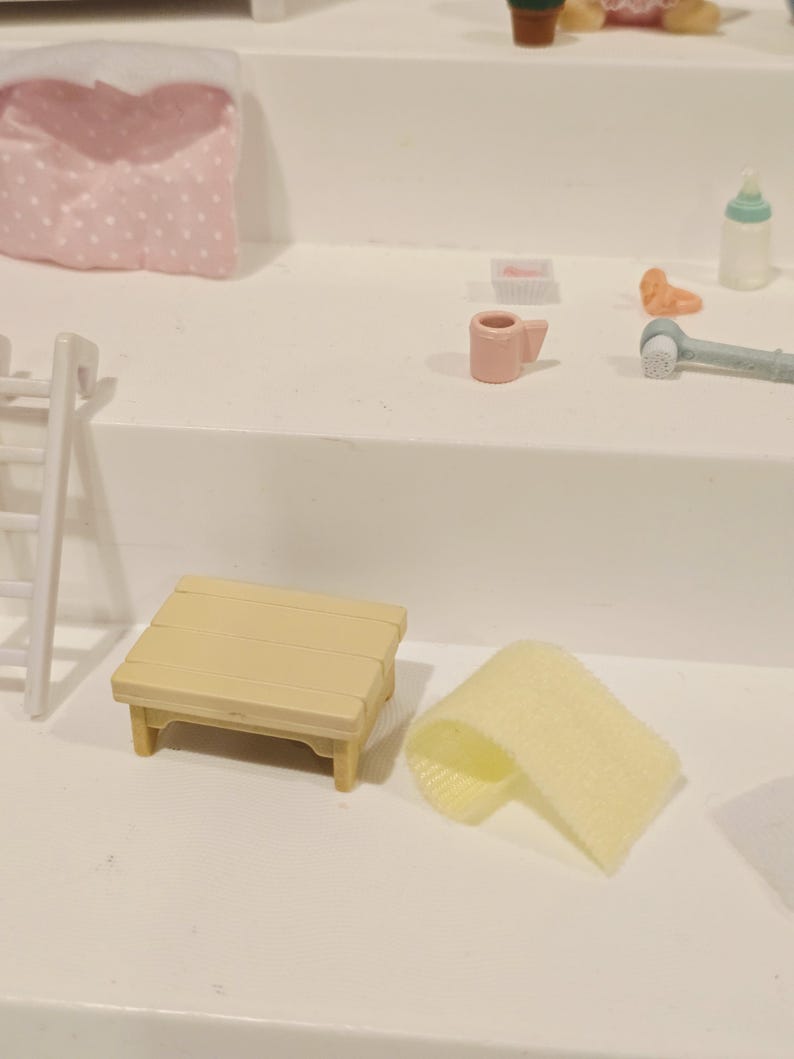 Puede incluir: Una escena en miniatura con una peque&ntilde;a mesa de picnic de color amarillo claro con una parte superior de listones y una toalla amarilla enrollada. Otros art&iacute;culos incluyen una taza rosa, un biber&oacute;n y una peque&ntilde;a almohada rosa con lunares blancos.