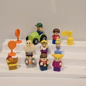 May include: A collection of colorful plastic toy figures and accessories, including a green tractor, a ghost, a chicken, and various characters. The toys are arranged on a white stepped surface, suggesting a playset or display.