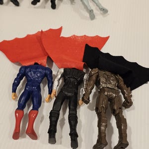 May include: A collection of superhero action figures, including Superman in a blue suit with a red cape, Thor in a black and silver outfit, and a brown armored figure. The figures are headless and arranged with their capes.