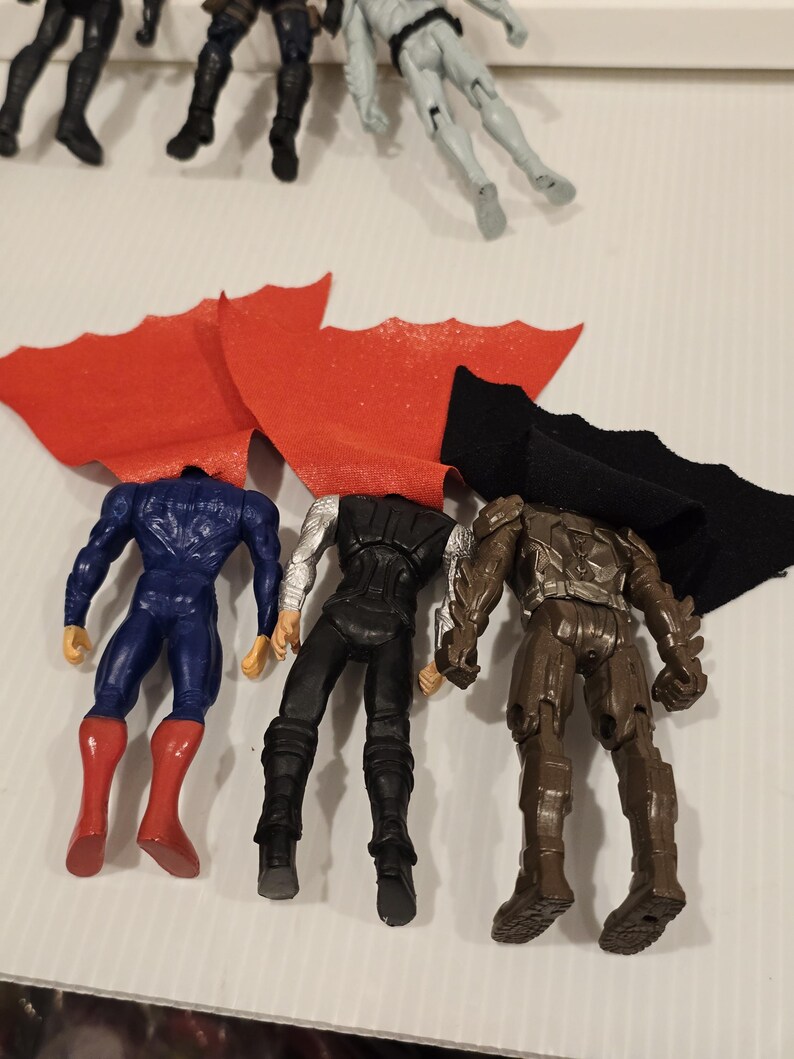 May include: A collection of superhero action figures, including Superman in a blue suit with a red cape, Thor in a black and silver outfit, and a brown armored figure. The figures are headless and arranged with their capes.