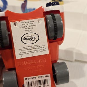 May include: A red toy car with gray wheels and a white and black top. The underside of the car has a Disney Store label with text. Other Stitch figurines are visible in the background.